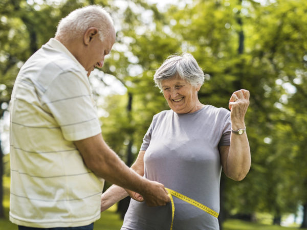 Senior man measuring his wife's waist with tape measure in the park. Focus is on woman.