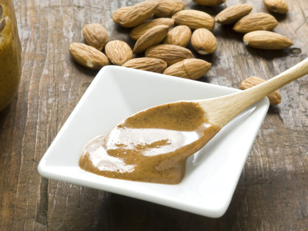A dish of homemade almond butter on rustic wood surface. Whole almonds in background and wooden spoon with fresh almond butter in foreground.